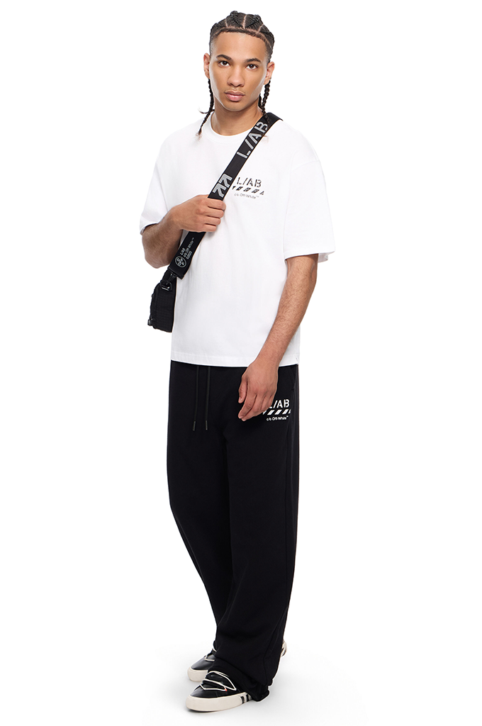 A man modeling a white crewneck t-shirt with black graphic text, black sweatpants with white text, and black sneakers, carrying a black shoulder bag with a white patterned strap against a white background.#color_white