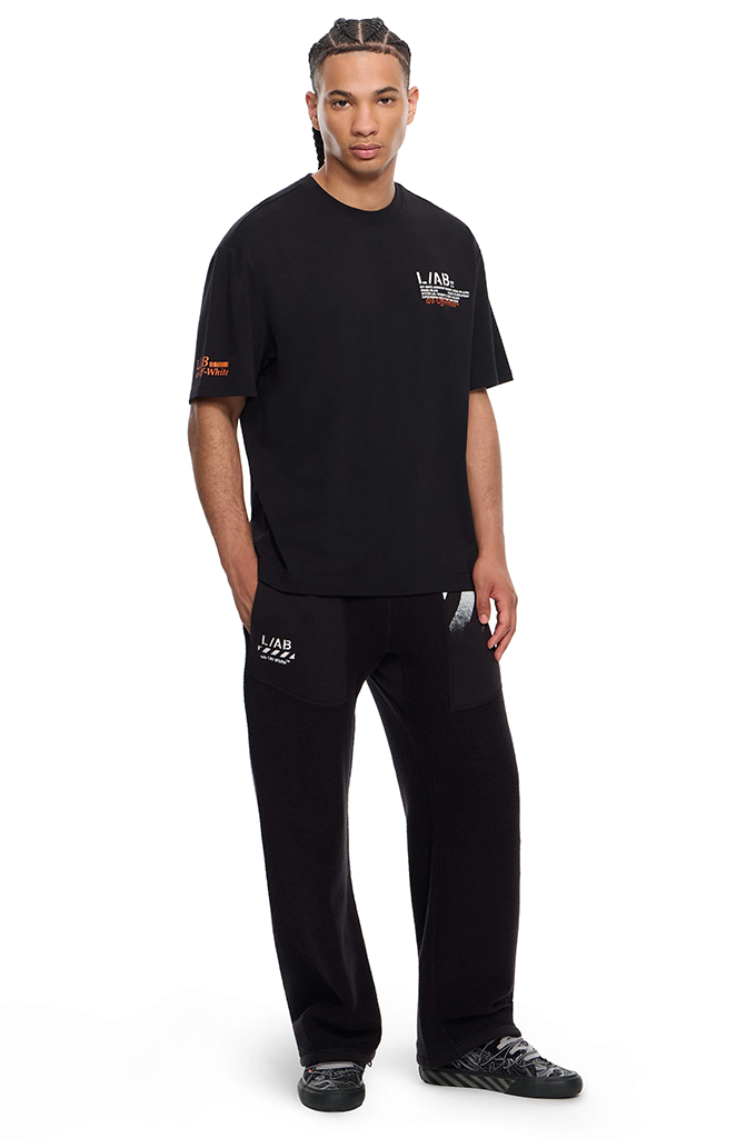 A man modeling a black short-sleeve t-shirt and matching black sweatpants set from LA x ZZZ, featuring small white and orange graphic details on the chest and sleeve, against a plain white background.#color_black