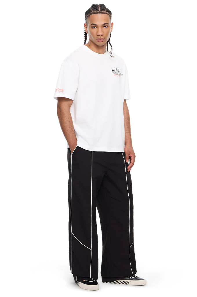 A model wearing a white short-sleeve t-shirt with a small black graphic on the left chest, paired with black wide-leg pants featuring white piping details, and black-and-white sneakers, against a plain white background.#color_white