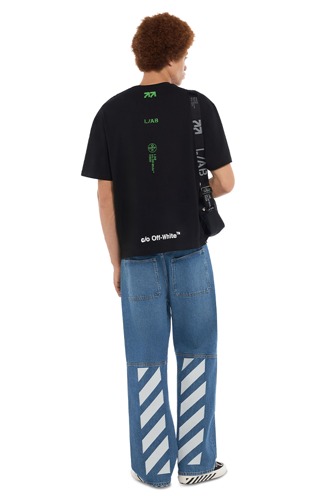 Back view of a male model wearing the L/AB c/o Off-White black Off Stacked cotton graphic t-shirt paired with Off-White blue diagonal stripe carpenter jeans and black sneakers.
