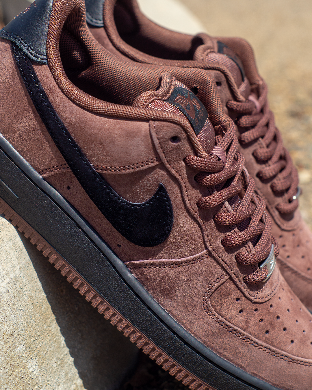 A close-up shot of a brown suede Nike Air Force 1 sneaker with a black Swoosh and black accents, resting on a textured stone surface.