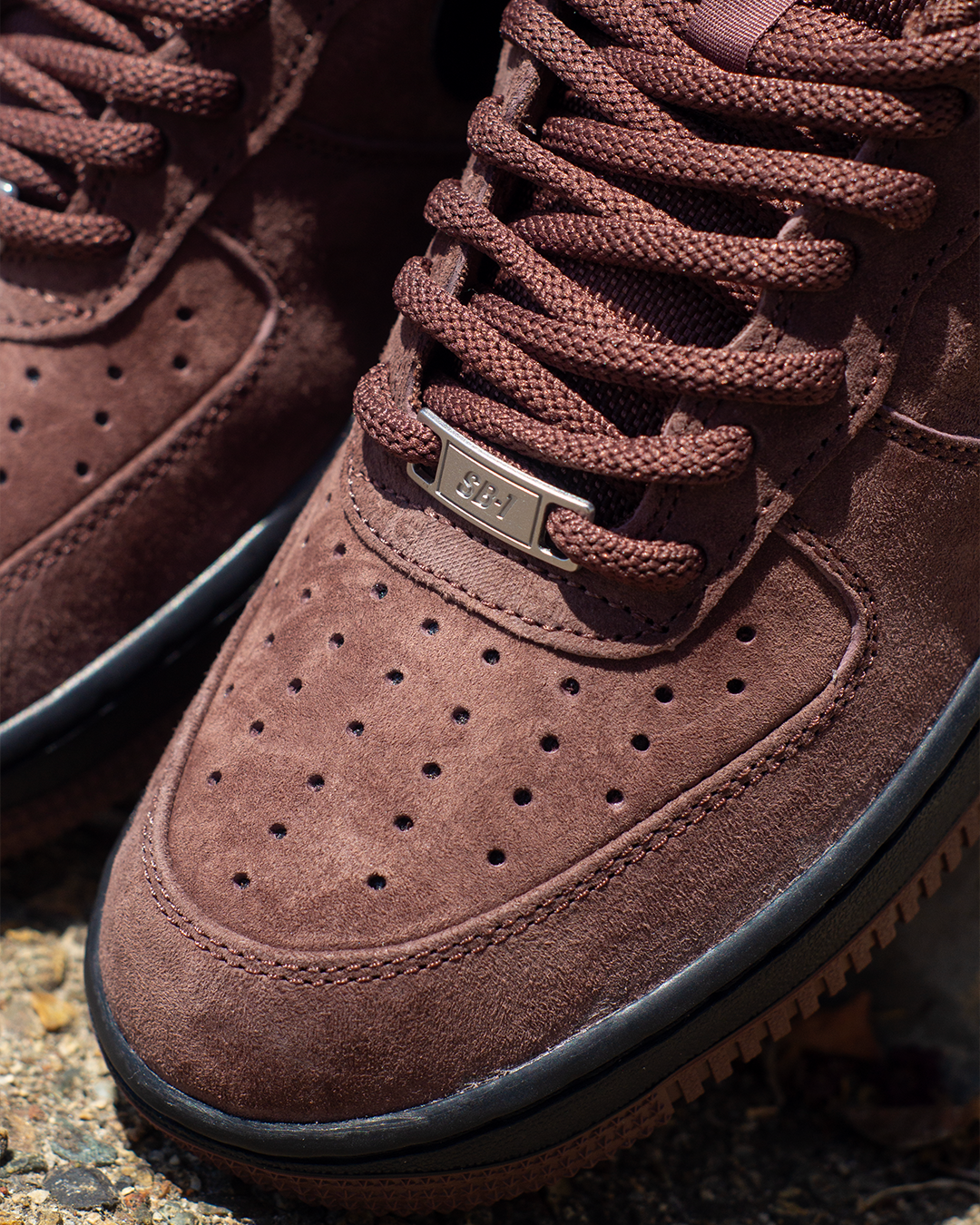 Close-up of a brown suede sneaker with matching brown laces, a small metallic logo plate, and a black rubber sole, photographed outdoors on a textured surface.