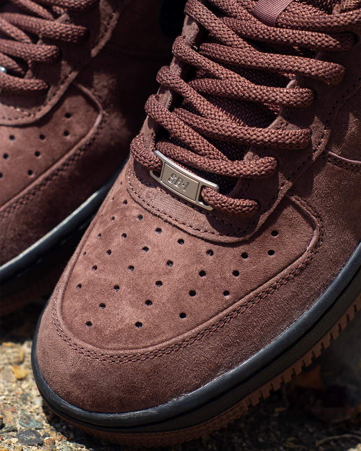Close-up of a brown suede sneaker with matching brown laces, a small metallic logo plate, and a black rubber sole, photographed outdoors on a textured surface.