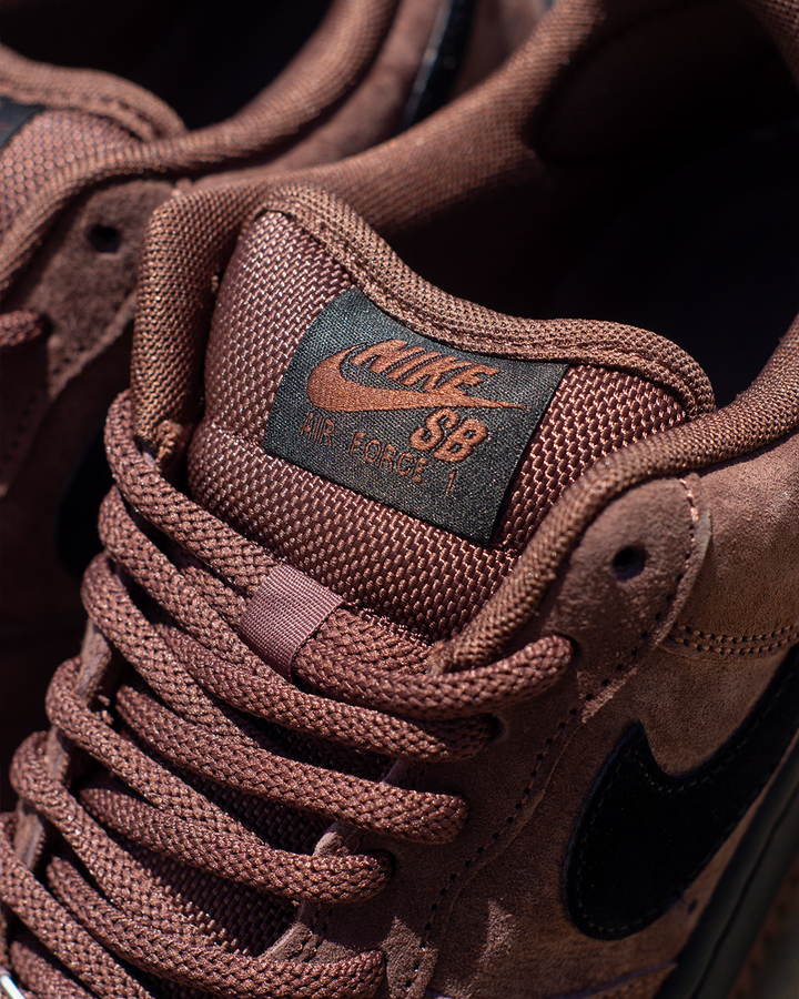 Close-up of a brown Nike SB sneaker showing the tongue label with 'Nike SB' and 'The Game 1' in orange, along with matching brown laces and a black Swoosh on the side.