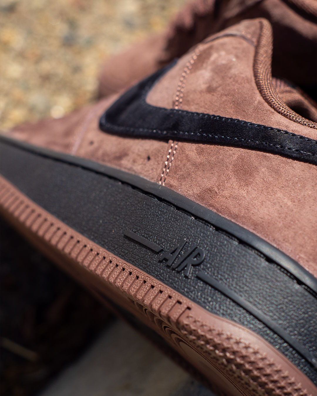 Close-up of a brown suede sneaker with a black Nike Swoosh and black midsole, showing the embossed 'AIR' text on the rubber sole.