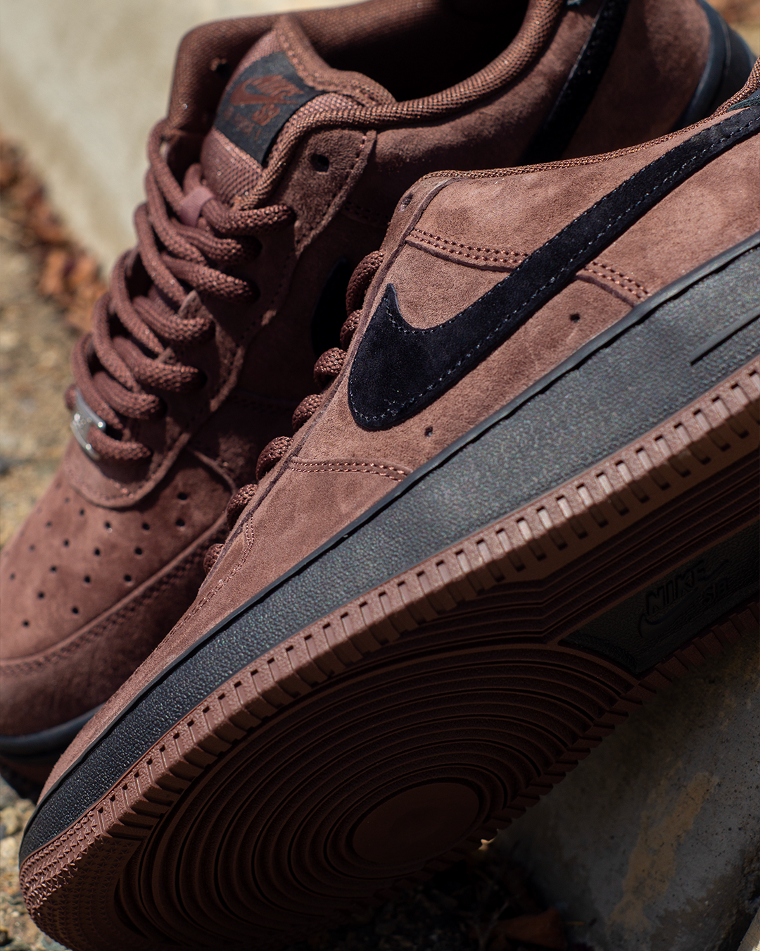 A close-up shot of a pair of brown suede Nike Air Force 1 Mid sneakers, showing the side profile with a black Swoosh and laces, resting on a stone surface.