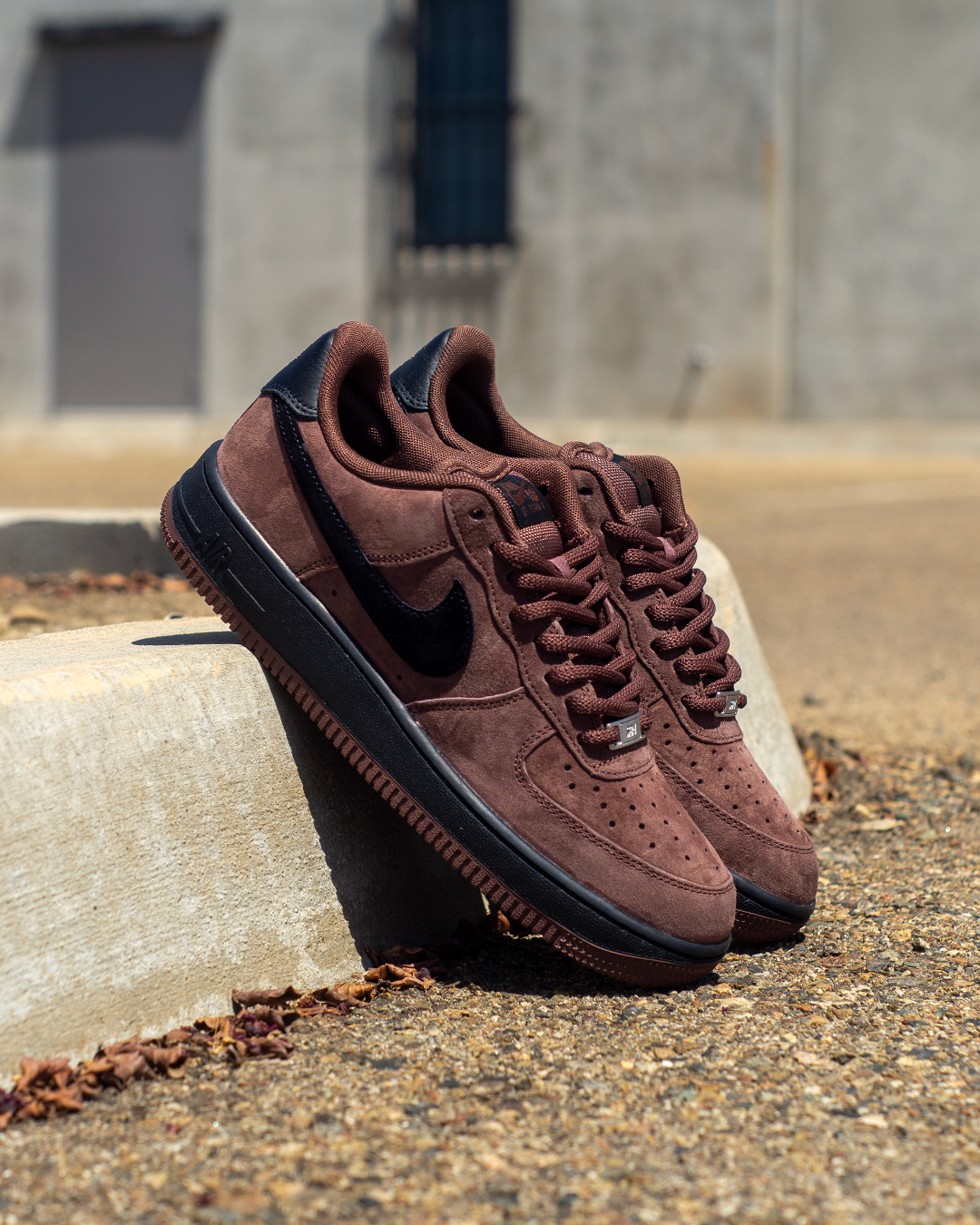 A pair of Nike Air Force 1 Low sneakers in brown suede and black, resting on a concrete curb outdoors.
