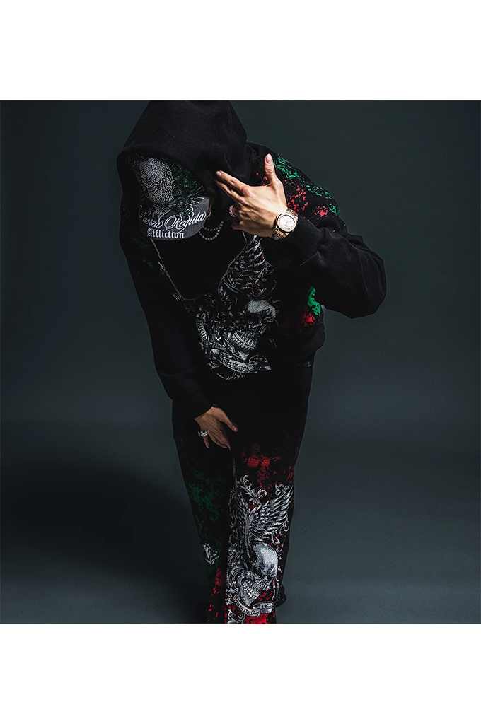 A person wearing a black hoodie and matching black pants with large white skull-and-bird graphics and red floral accents, posed against a dark studio background.