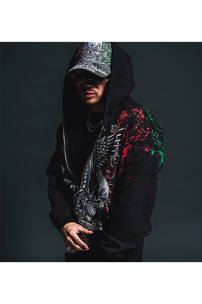 A man wearing a black hoodie with a large black-and-white eagle graphic on the front and red and green paint-splatter details on the right sleeve, posed against a dark studio background.