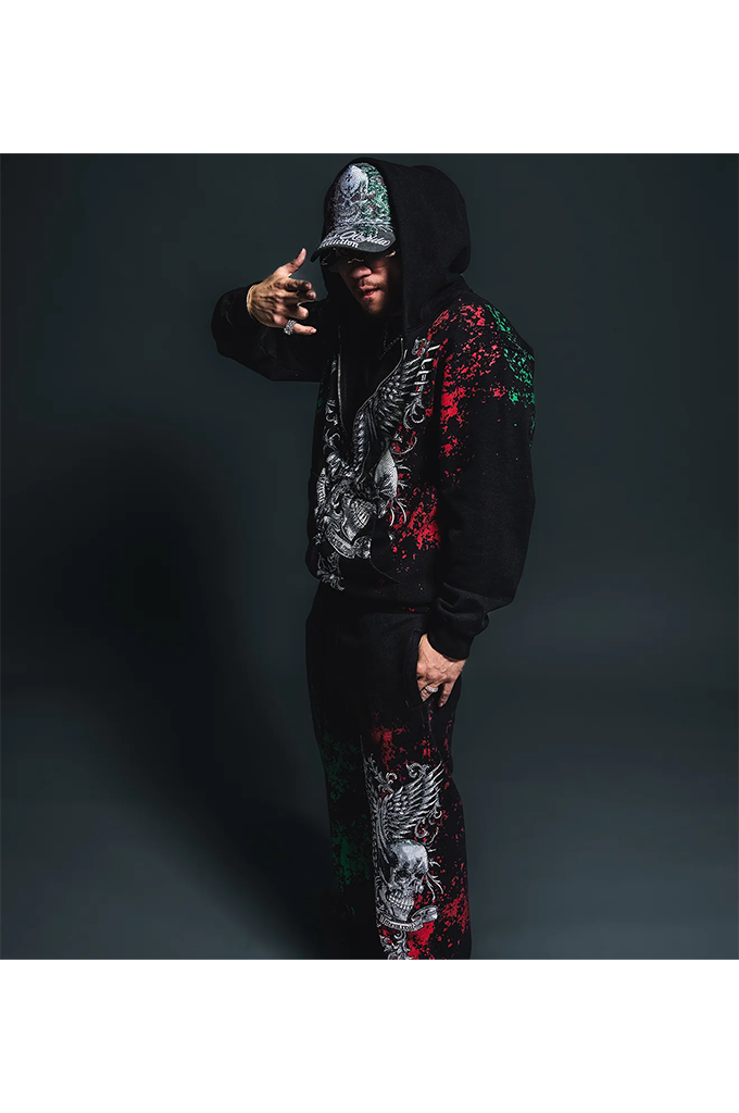 A man wearing a black hooded tracksuit with red and green graphic print details, posing against a dark studio background.