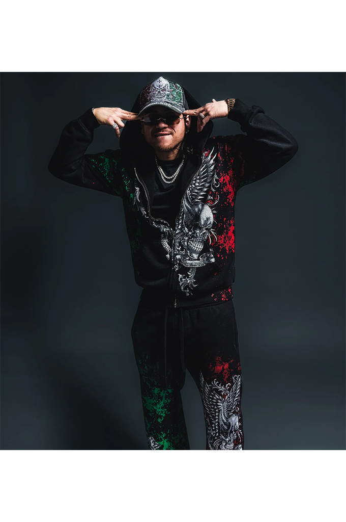 A man modeling a black graphic tracksuit featuring a large eagle design in white and red, with green splatter accents on the sleeves and pants, set against a dark studio background.