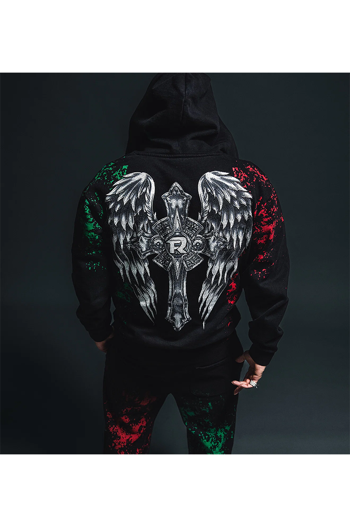 A person shown from the back wearing a black hoodie with a large winged cross graphic in white, silver, and red on the back, paired with matching black pants with red and green splatter details, against a dark studio background.