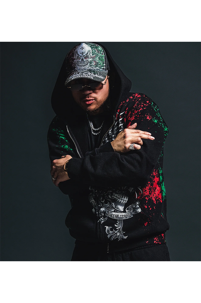 A man wearing a black hoodie with red and green paint splatter graphics, a silver graphic print on the front, and a camouflage-patterned baseball cap, posed against a dark studio background.