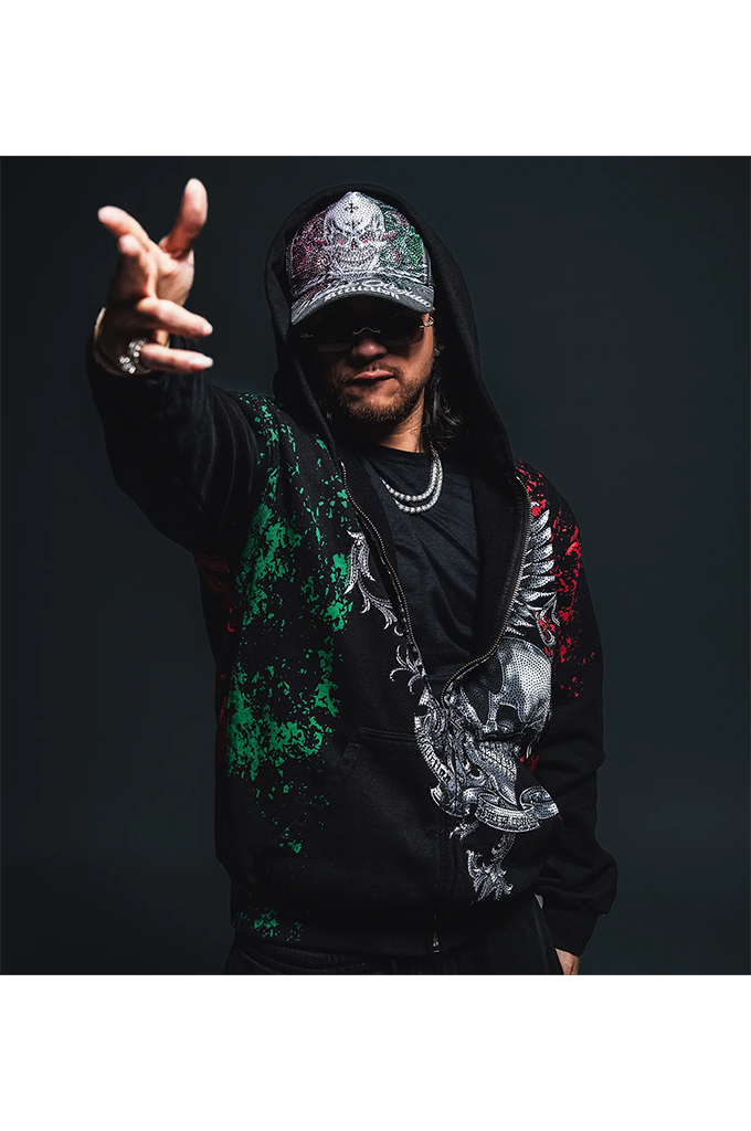 A man wearing a black hoodie with green and red paint splatter details, a silver skull graphic on the front, a silver chain necklace, and a silver cap, posing with one hand raised against a dark background.