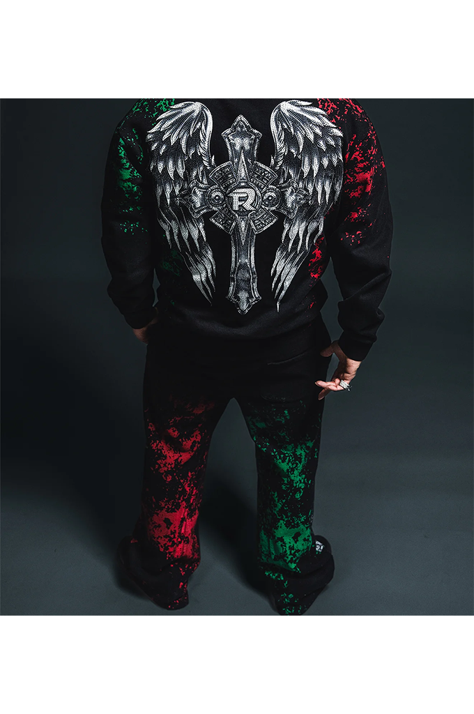A person shown from the back wearing a black hoodie and matching sweatpants with a large white angel-wing and cross graphic on the back, plus red and green splatter accents on the sleeves and legs, against a dark studio background.