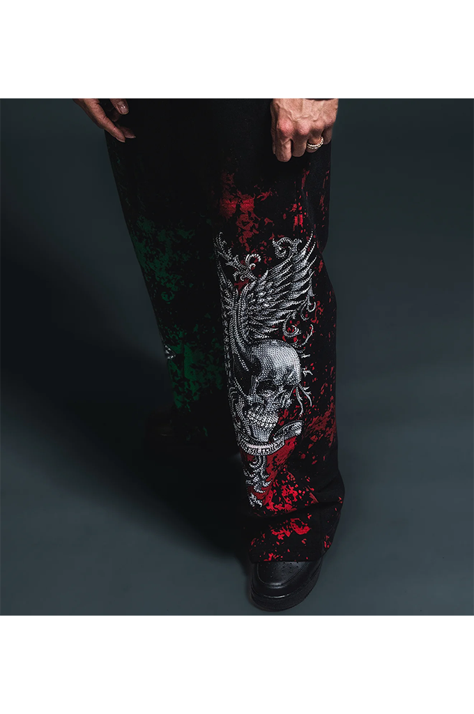 Black pants featuring a large white skull and wing graphic, with red and green distressed splatter accents, shown on a model against a dark studio background.