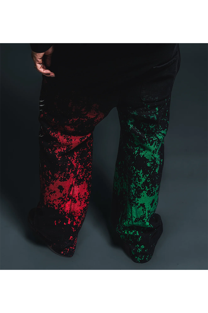A person wearing loose black pants with a high-contrast red and green splatter pattern on the lower legs, shown from the waist down against a dark studio background.