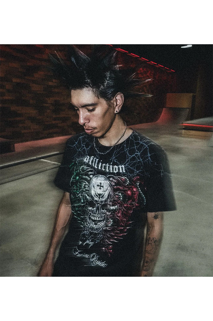 A man with a high-spiking mohawk wears a black graphic t-shirt featuring the word 'affliction' and a large skull illustration with red, green, and white details. The photo is taken in a dimly lit indoor space with concrete floors and brick walls.