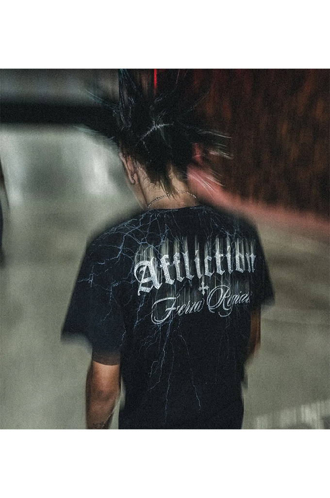A person shown from behind wearing a black graphic t-shirt with 'Addiction From Regime' printed in white gothic and script fonts on the back, set against a dark, moody background.