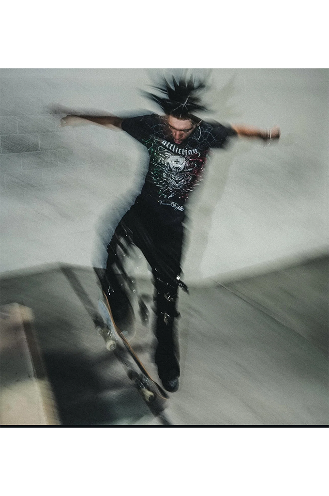 A person in a black graphic t-shirt and black pants skateboarding on a concrete surface, captured with motion blur and a dramatic spiked hair effect against a gray background.