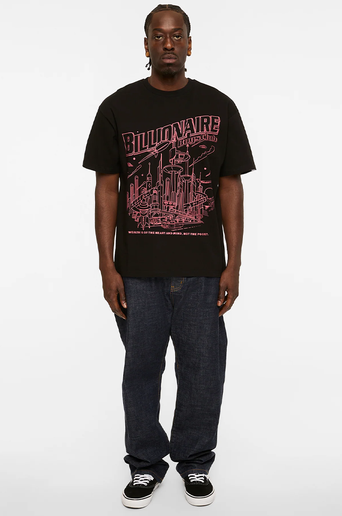 A man modeling a black t-shirt with a pink graphic print that reads 'Billionaire Boys' on the front, paired with dark blue jeans and black sneakers, against a plain white studio background.#color_black