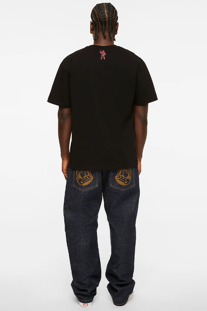A man viewed from behind wearing a black t-shirt with a small pink graphic on the back neck, dark blue jeans with gold embroidery on the back pockets, and white sneakers, set against a plain white studio background.#color_black
