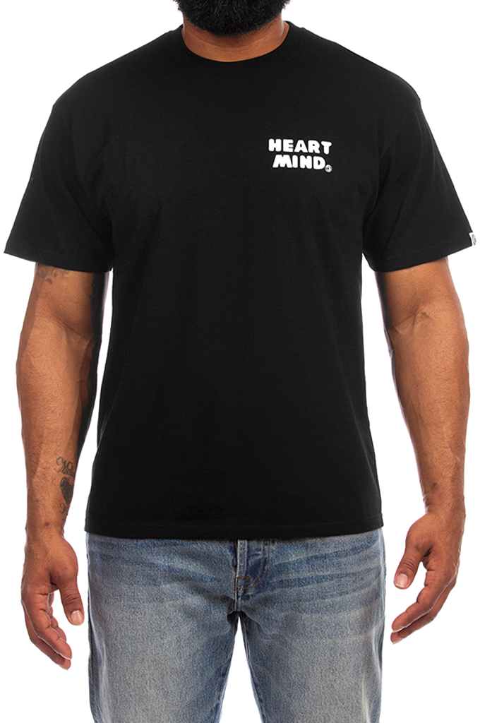 A man wearing a black short-sleeve t-shirt with 'HEART MIND.' printed in white text on the left chest, paired with blue jeans against a plain white background.