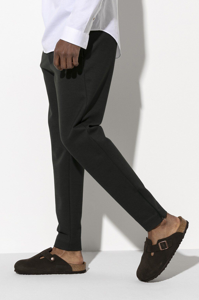 A person wearing slim black trousers and dark brown clog-style mules, cropped from the waist down, walking against a white wall with a strong shadow.#color_mocca