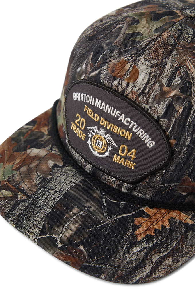 Close up of a medium-profile Brixton snapback hat featuring an all-over tree camo pattern. The cap has a flat bill with a black accent rope across the brim, a green-brown camouflage crown, and a central black embroidered patch that reads "Brixton Manufacturing Field Division 2004".
