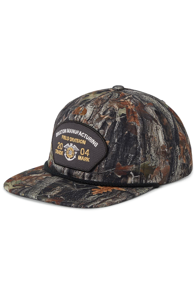 A medium-profile Brixton snapback hat featuring an all-over tree camo pattern. The cap has a flat bill with a black accent rope across the brim, a green-brown camouflage crown, and a central black embroidered patch that reads "Brixton Manufacturing Field Division 2004".
