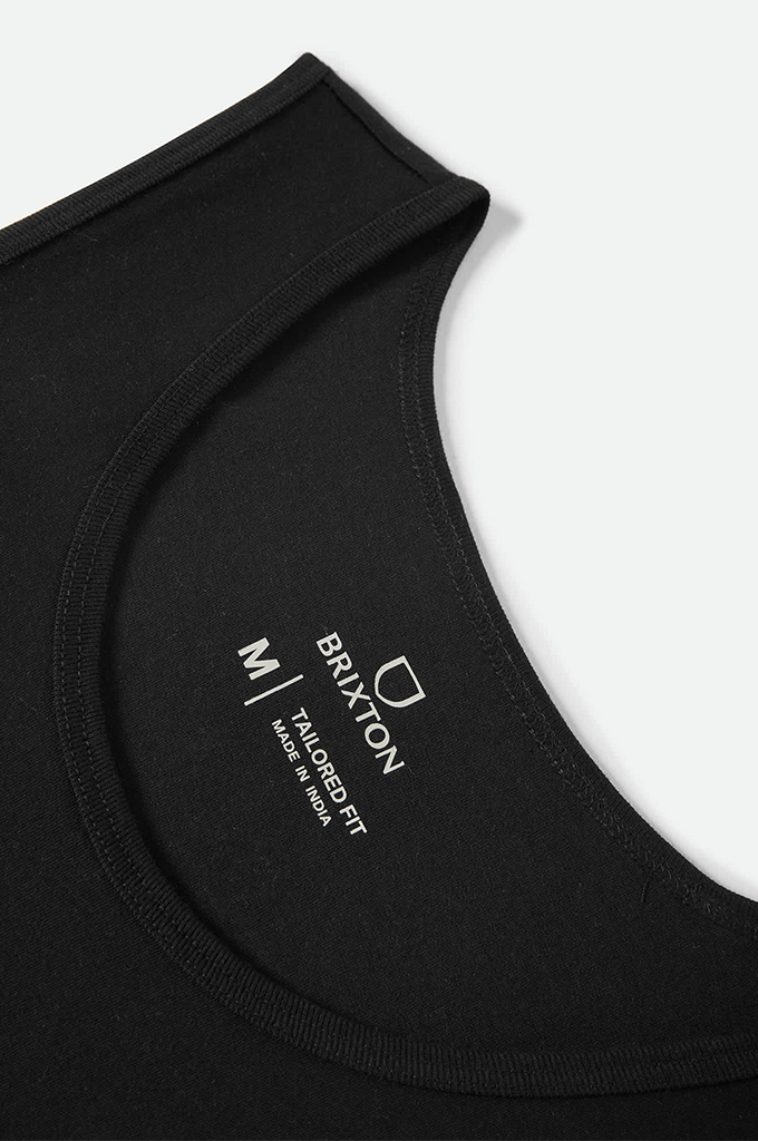 A close-up shot of the neckline of a black garment, showcasing the inner neck label printed in white text. The label features a shield-shaped logo above the brand name "BRIXTON". Below the brand name, it reads "TAILORED FIT" and "MADE IN INDIA". To the left of a vertical separating line, the letter "M" indicates the size. The fabric has a smooth texture with clean stitching along the collar trim.