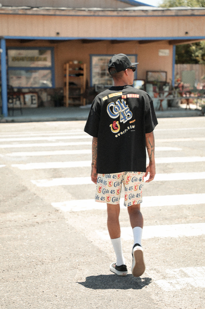 An image of a person seen from behind walking outdoors on a sunny day. They are wearing a black oversized graphic t-shirt featuring a colorful "Colt 45" logo and horseshoe design on the back, paired with cream-colored shorts covered in an all-over repeating "Colt 45" pattern. They complete the outfit with a black baseball cap worn backward, high white socks, and dark slip-on shoes.
