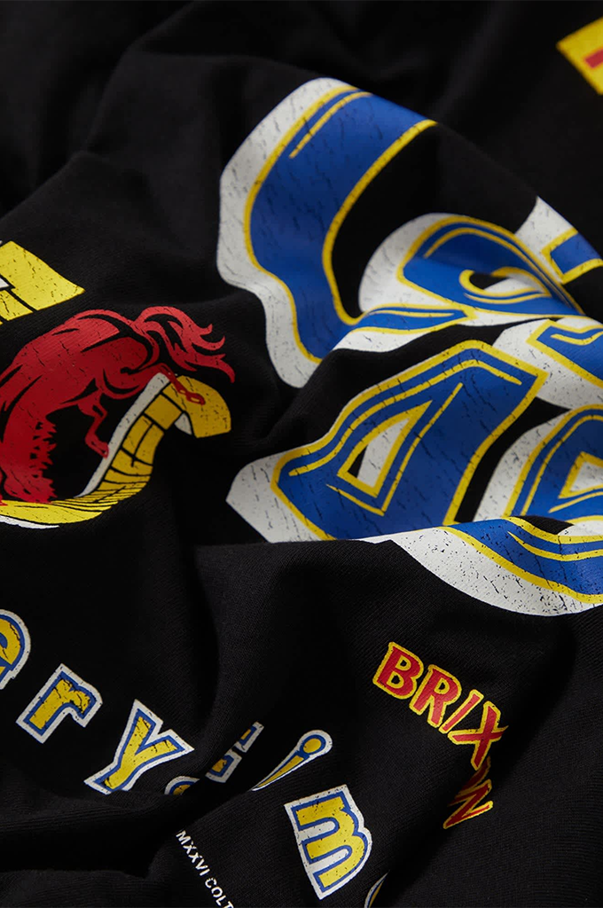 Close-up of a black folded fabric featuring a vintage streetwear graphic design. The print showcases a large stylized blue and white typography logo outlined in bright yellow, alongside a red and gold kicking mule emblem. The orange and red text "BRIXTON" is visible near the bottom right corner.
