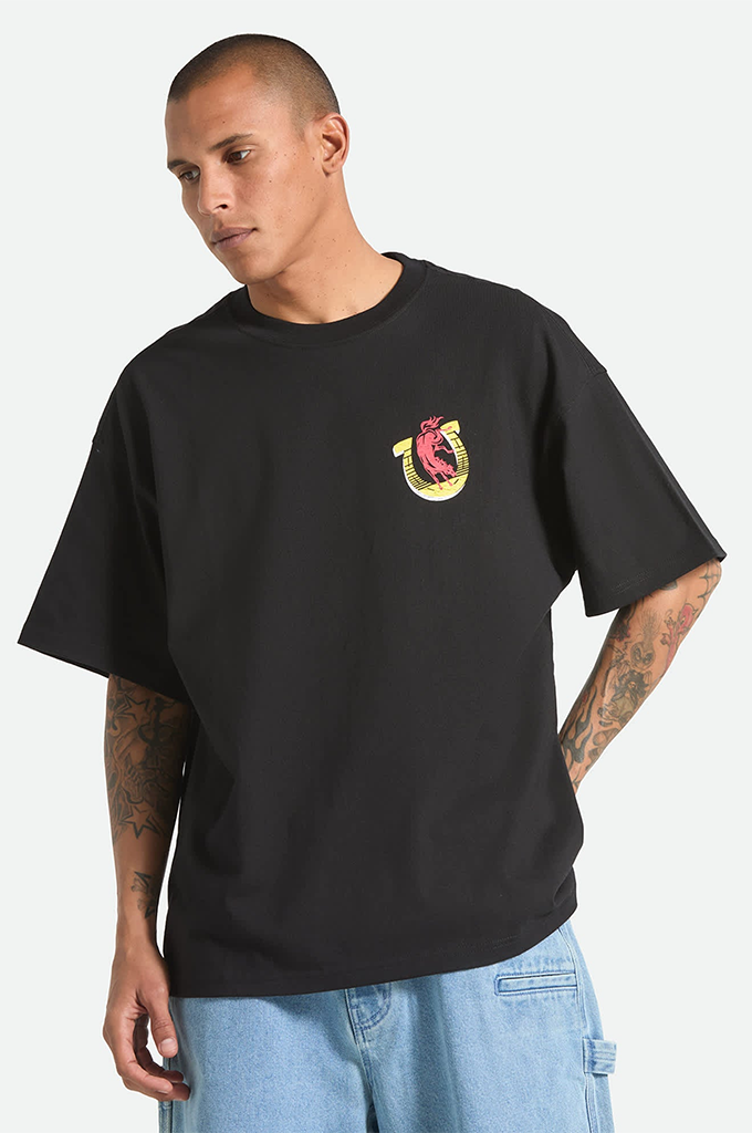 A young man with a shaved head and tattoos on his arms is modeling a black, oversized Brixton x Colt 45 graphic t-shirt against a plain white background. The short-sleeve shirt has a relaxed fit with dropped shoulders and features a small logo on the left chest depicting a yellow horseshoe with a pink bull inside. He is also wearing light blue denim carpenter shorts, shown from the waist down.