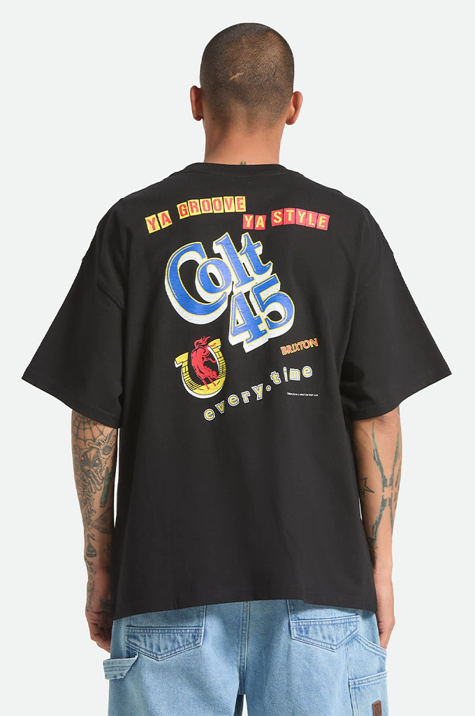 A young man with a shaved head and tattoos on his arms is modeling a black, oversized Brixton x Colt 45 graphic t-shirt against a plain white background. The short-sleeve shirt has a relaxed fit with dropped shoulders and features a large logo on the back depicting a yellow horseshoe with a pink bull inside. He is also wearing light blue denim carpenter shorts, shown from the waist down.