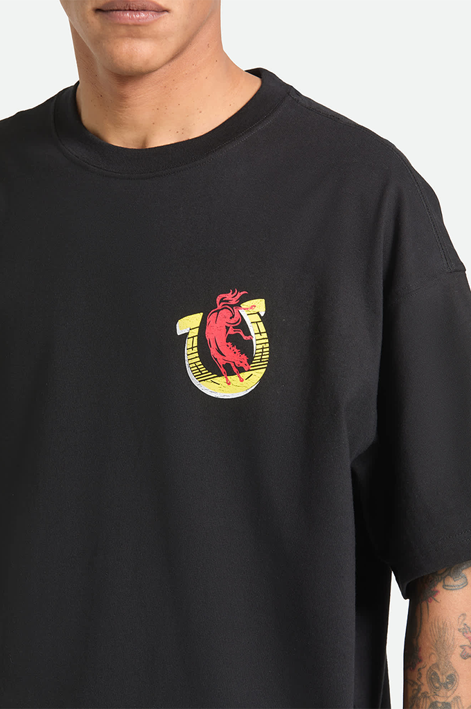 A young man with tattoos on his arms is modeling a black, oversized Brixton x Colt 45 graphic t-shirt against a plain white background. The short-sleeve shirt has a relaxed fit with dropped shoulders and features a small logo on the left chest depicting a yellow horseshoe with a pink bull inside. 