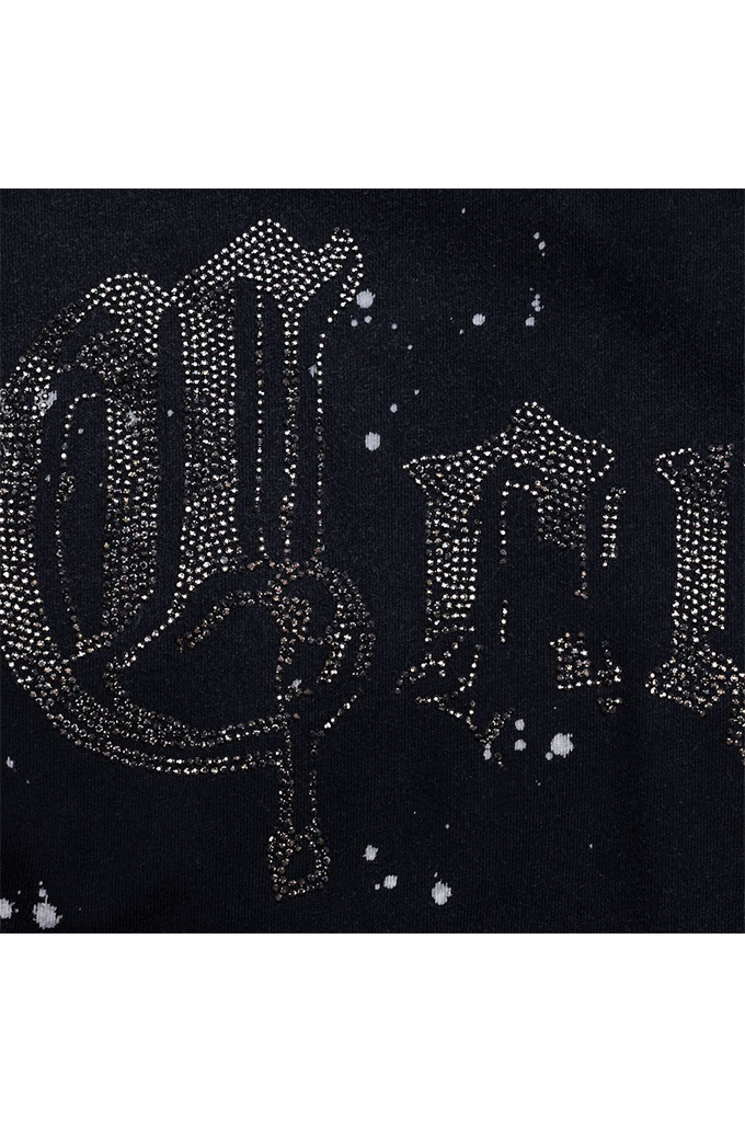 Close-up of a black garment featuring a large gothic-style letter 'C' embellished with rhinestones, alongside a distressed 'F' and scattered white paint splatter details.