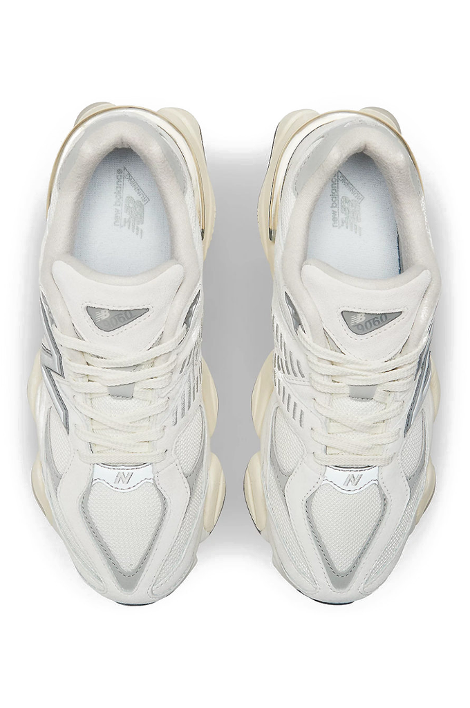 A top-down view of a pair of chunky, cream-colored New Balance 9060 lifestyle sneakers. The shoes feature a mix of off-white mesh underlays and suede overlays with metallic silver details on the toe and sides. They are designed with a distinctly sculpted, wavy midsole and white laces, set against a plain, light grey background. #color_white-grey-sea-salt-concrete