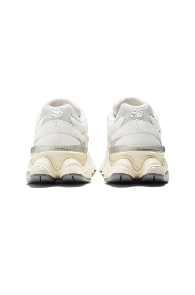 A back view of a New Balance 9060 sneaker. The shoe features a retro-futuristic style with a chunky, sculpted cream and grey midsole. The upper is made of off-white mesh and suede overlays, with a metallic silver "N" logo on the side. #color_white-grey-sea-salt-concrete