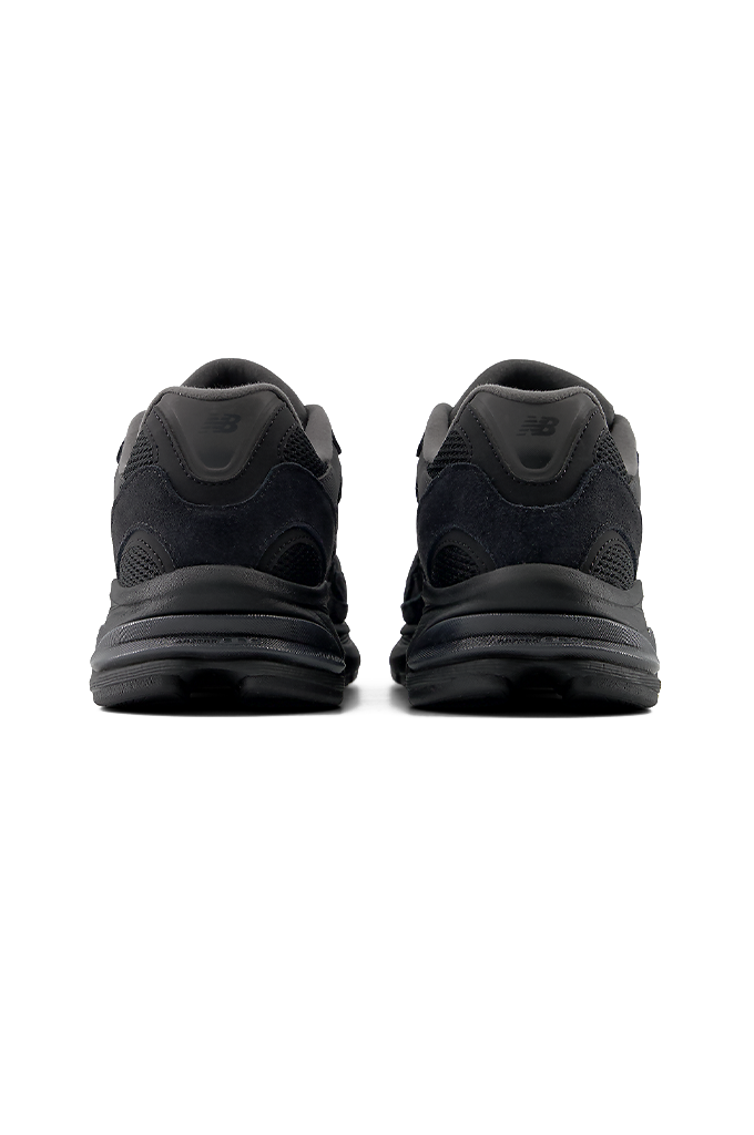 A back view of a New Balance 2010 sneaker in an all-black colorway. The running-style shoe features a dark mesh base with suede overlays, matching black laces, and the signature New Balance "N" logo on the side panel. The entire sneaker, including the thick, segmented midsole and rubber outsole, is uniformly black.#color_black-black