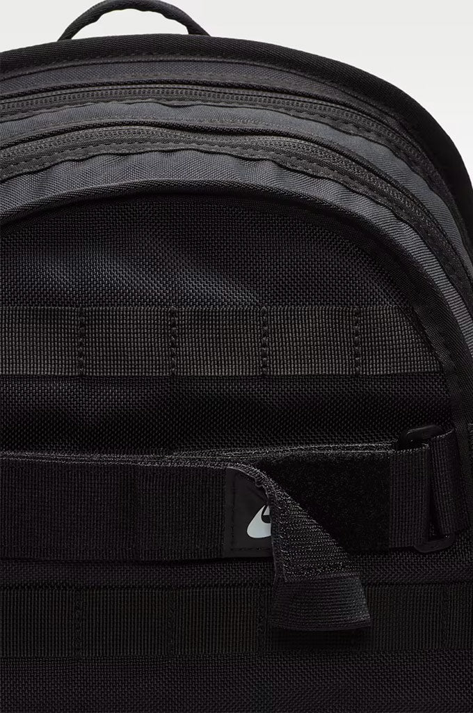 Close-up of a black backpack showing the front zippered pocket with a small white Nike swoosh logo on a black tab, set against a light gray background.#color_black