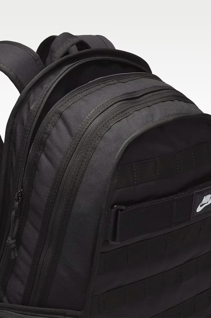 A close-up of a black Nike backpack showing the front pocket with a white Nike swoosh logo, set against a light gray background.#color_black