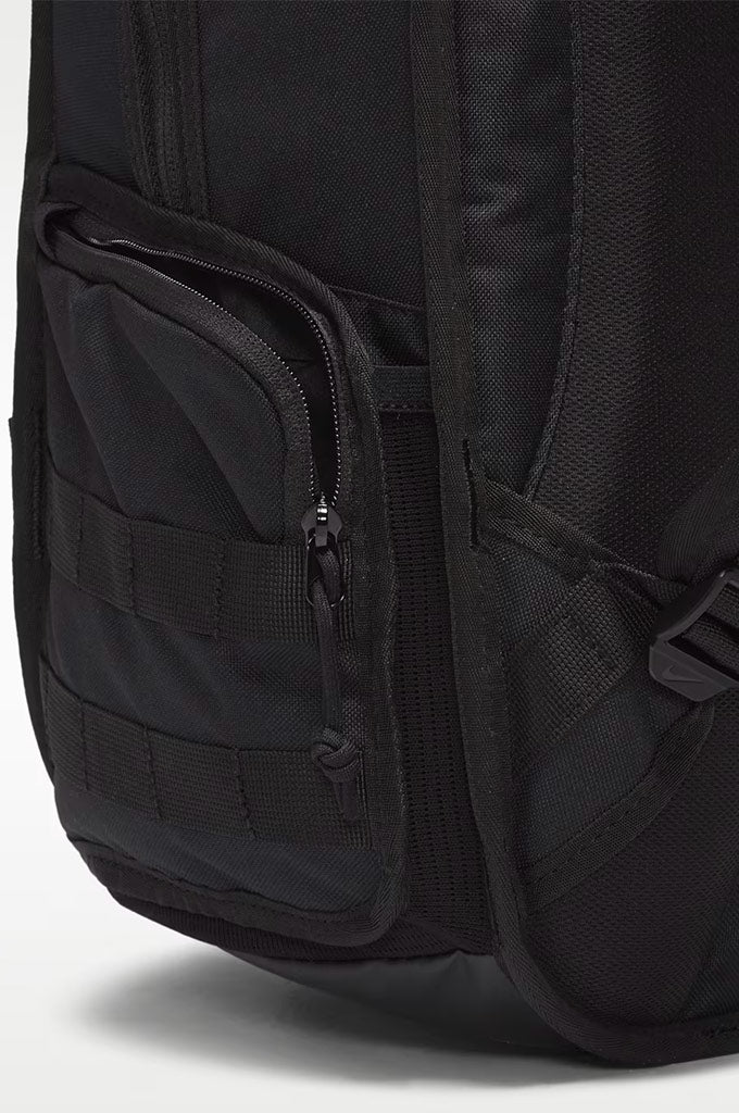 Close-up of a black backpack showing a front zippered pocket with webbing and side compression straps on a light gray background.#color_black