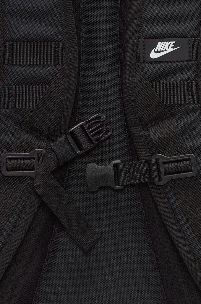 Close-up of a black Nike backpack showing the padded shoulder straps, buckle closures, and a white Nike logo patch on the upper right side.#color_black