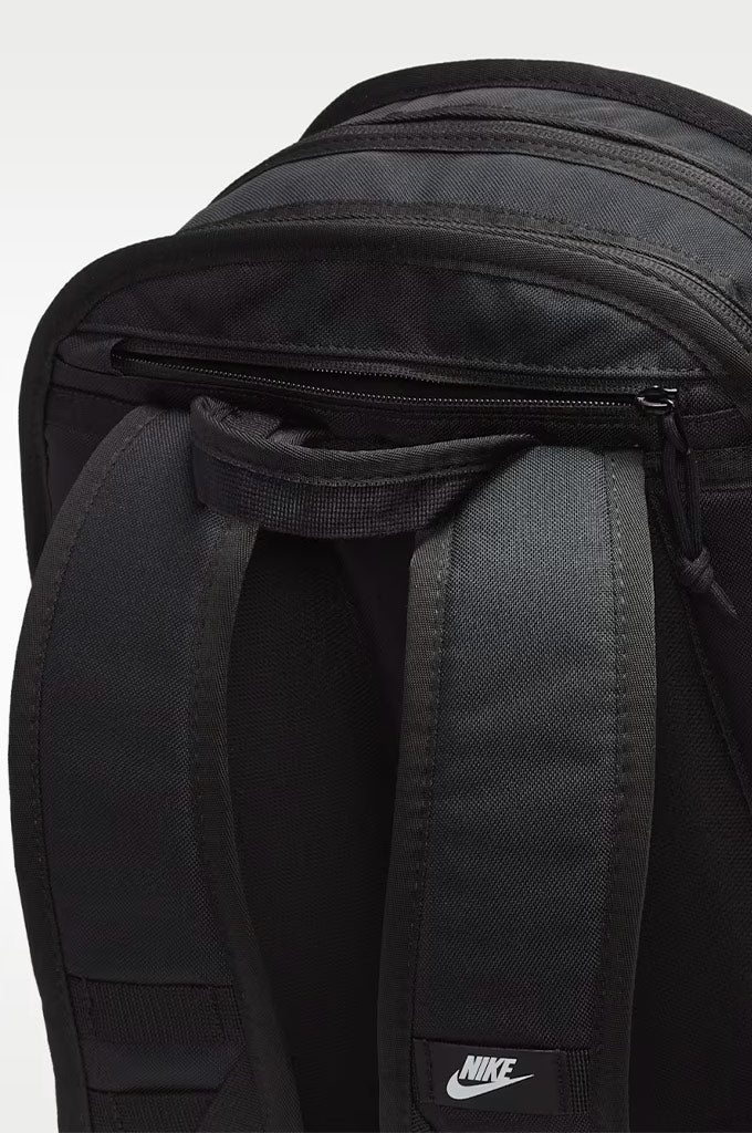 Close-up of a black Nike backpack showing the padded shoulder straps, zipper compartment, and a small white Nike logo patch near the bottom.#color_black