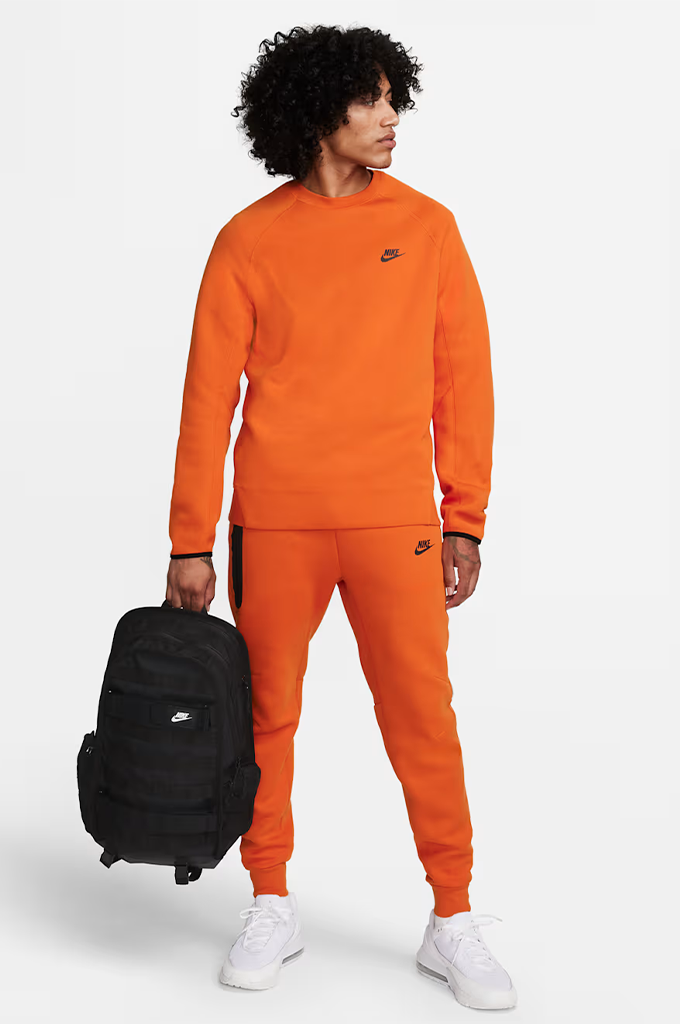 A man modeling a bright orange Nike tracksuit, consisting of a crewneck sweatshirt and matching jogger pants, while holding a black backpack against a light gray studio background.#color_black