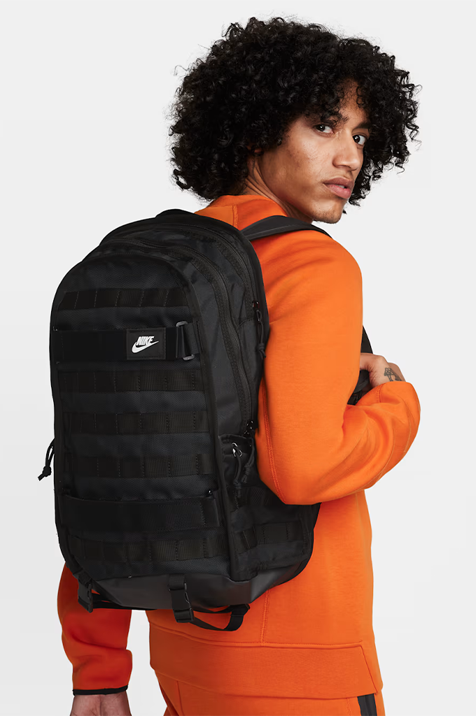A man wearing a bright orange sweatshirt and black Nike backpack, photographed from the back against a light gray studio #color_black