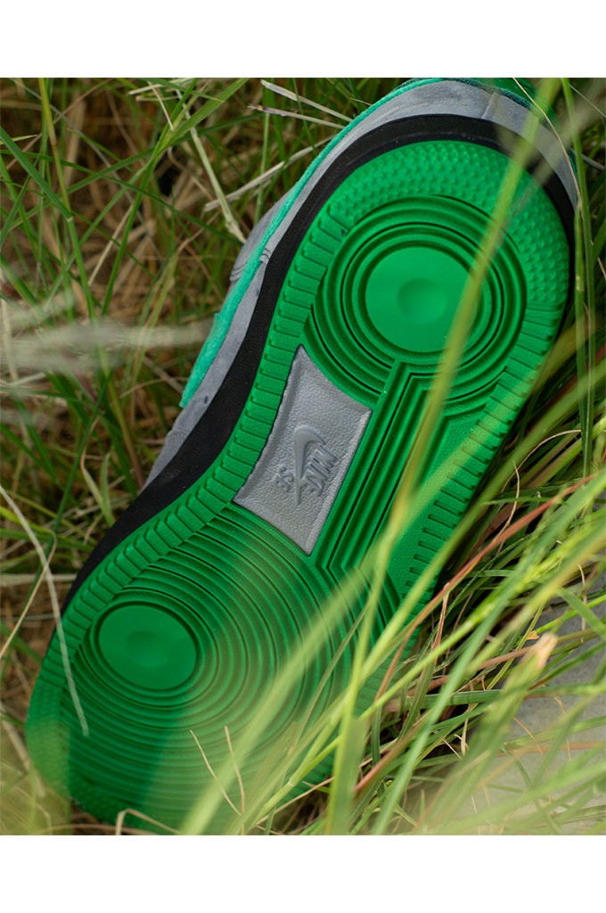 Bottom sole view of Nocturnal x Nike SB Air Force 1 Low SE sneaker in Cool Grey and Court Green, showcasing the translucent green rubber traction pattern and Nike SB logo amidst green grass.
