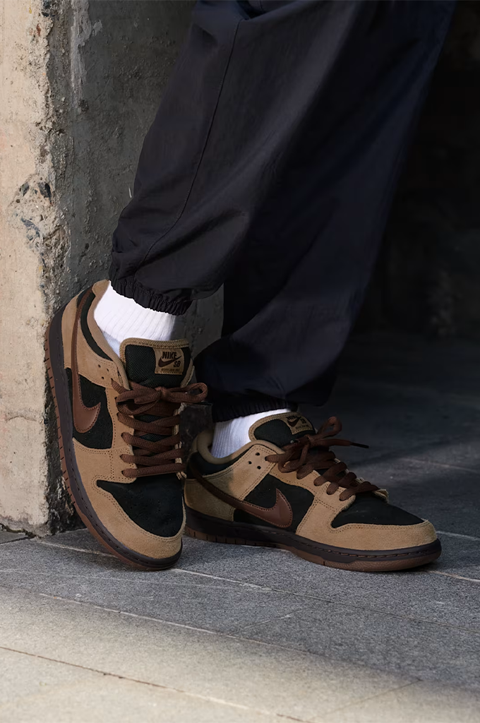 Casual view of a Nike Dunk sneaker in a colorway of tan suede, black leather, and dark brown. The shoe features a tan suede upper, black leather overlays, dark brown laces, a dark brown Swoosh, and a dark brown gum sole, shown from a side profile view on a white background.