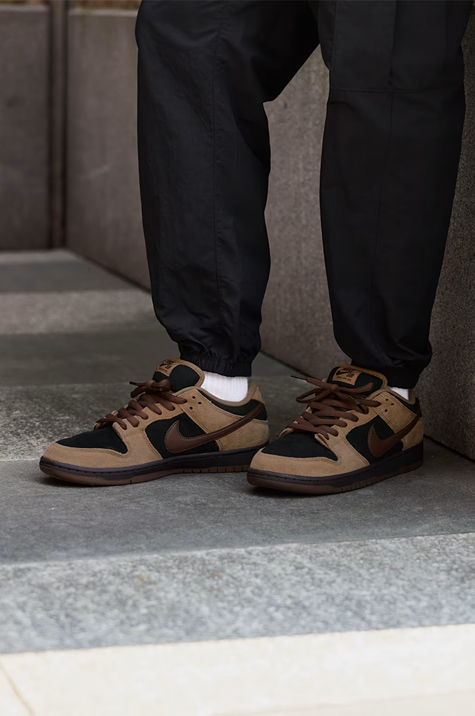 Casual view of a Nike Dunk sneaker in a colorway of tan suede, black leather, and dark brown. The shoe features a tan suede upper, black leather overlays, dark brown laces, a dark brown Swoosh, and a dark brown gum sole, shown from a side profile view on a white background.